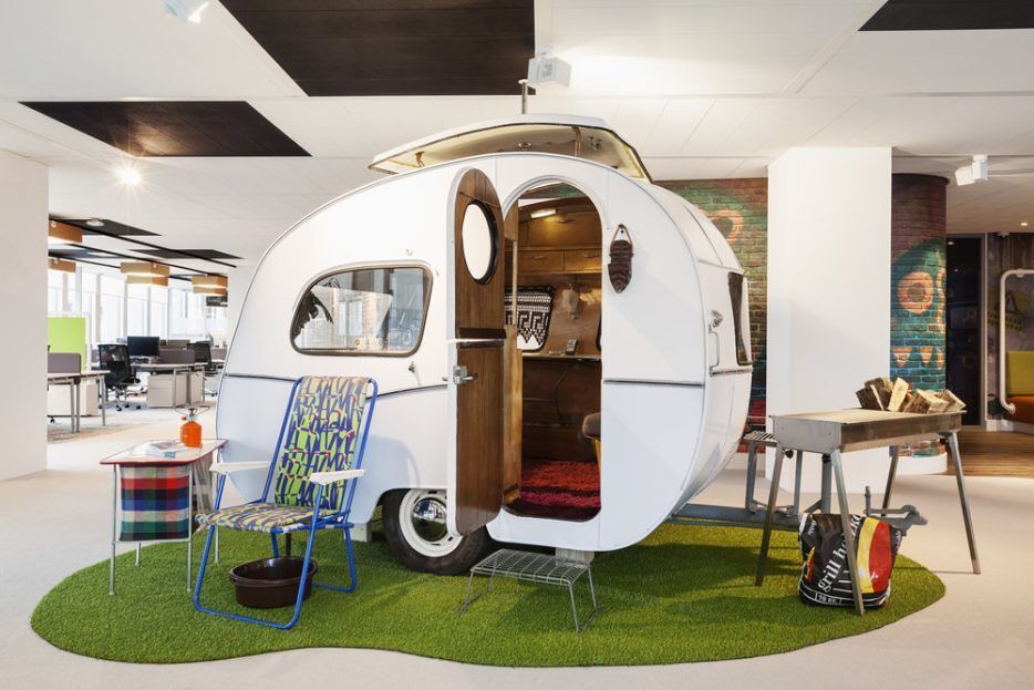 Petite caravane blanche sur un tapis de fausse herbe au centre d'un open-space, table et chaise de camping devant et barbecue à droite