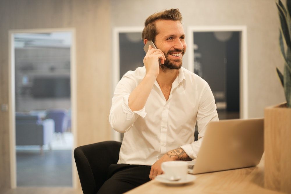 Un homme en chemise blanche est assis à son bureau. Il est au téléphone et un ordinateur portable et une tasse de café sont posés devant lui