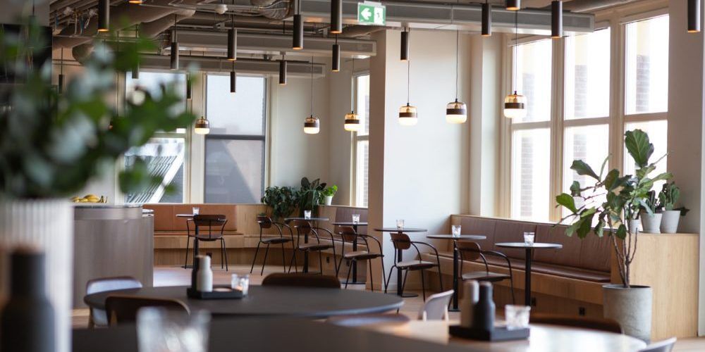 La salle d'une cafétéria dans les tons bois. Des banquettes sont sur le long des murs et fenêtres avec des petites tables rondes ainsi que des chaises en bois et métal noir