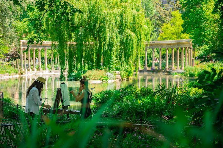 Au premier plan, des feuilles de plante floues. Au second plan, deux personnes entrain de peindre devant un étang avec des colonnes en fond. Tout l'étang est entouré de verdure