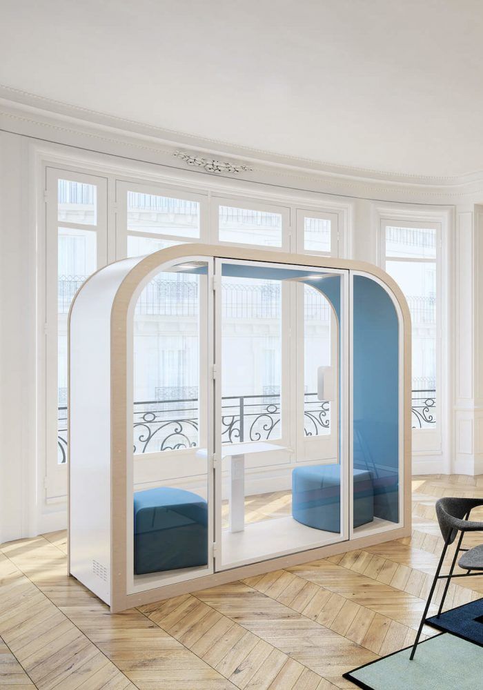 Phonebox blanche aux coins supérieurs arrondis, aux parois vitrées et bleues contenant deux sièges bleux et une petite table blanche placée devant de grandes fenêtres sur un parquet clair