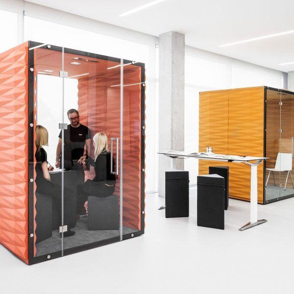 Deux phoneboxes, l'une rose, l'autre orange, avec des parois vitrées et trois personnes discutant à l'intérieurs sur des tabourets gris 