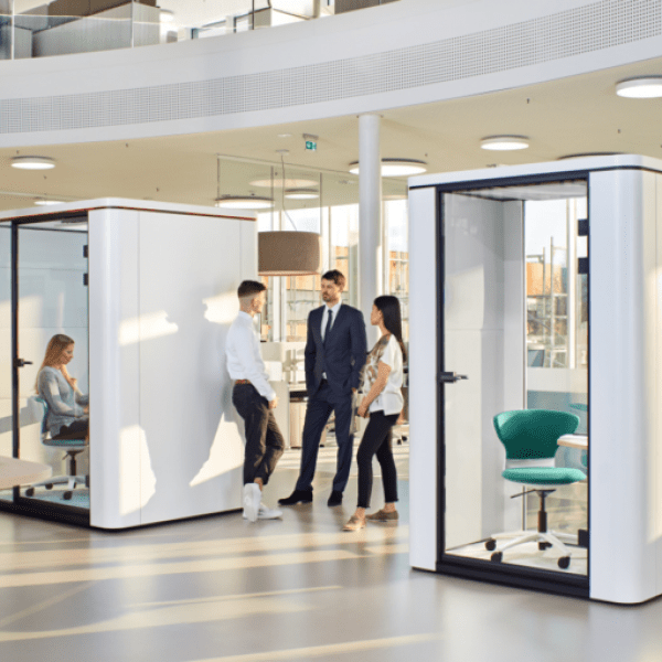 Trois personnes discutant entre deux phoneboxes se trouvant dans un open-space, les phoneboxes sont blanche avec un siège vert et un bureau à l'intérieur